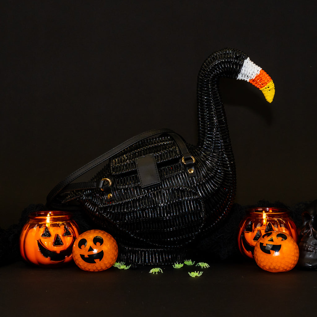 A flamingo shaped wicker handbag, painted shiny black over the body, with a white, orange and yellow candycorn themed beak. It has two black leather straps and a black leather fastening. 