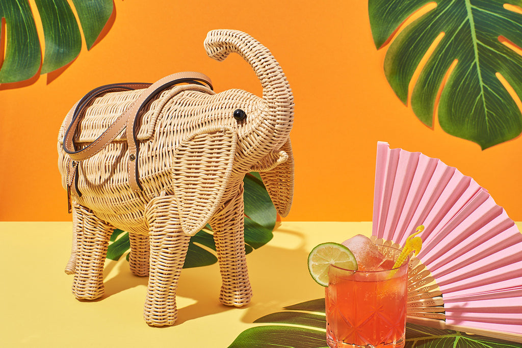 The natural Wicker coloured Belle-ephant the Elephant handbag sits in front of an orange, yellow and monsterra leaf covered background. With leather handles and a raised trunk, the retro inspired handbag is perfect for adventures of bold fashionistas.