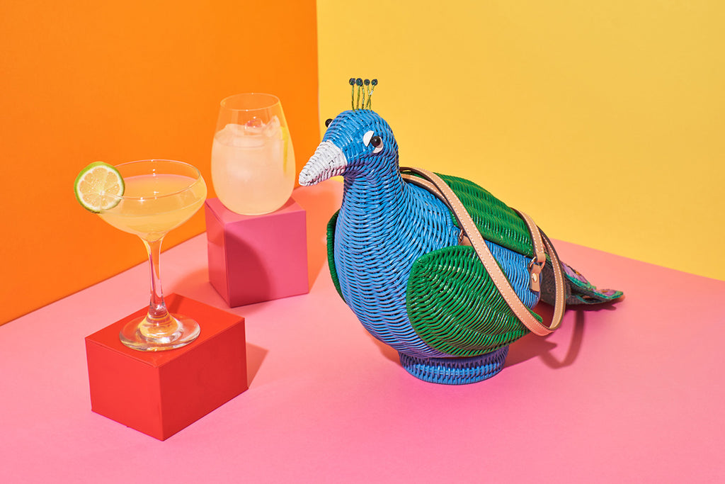 A peacock shaped handbag by Australian label Wicker Darling sits in a colourful background surrounded be cocktails. The bag is mostly blue with green wings, beaded crest and an embroidered tail.