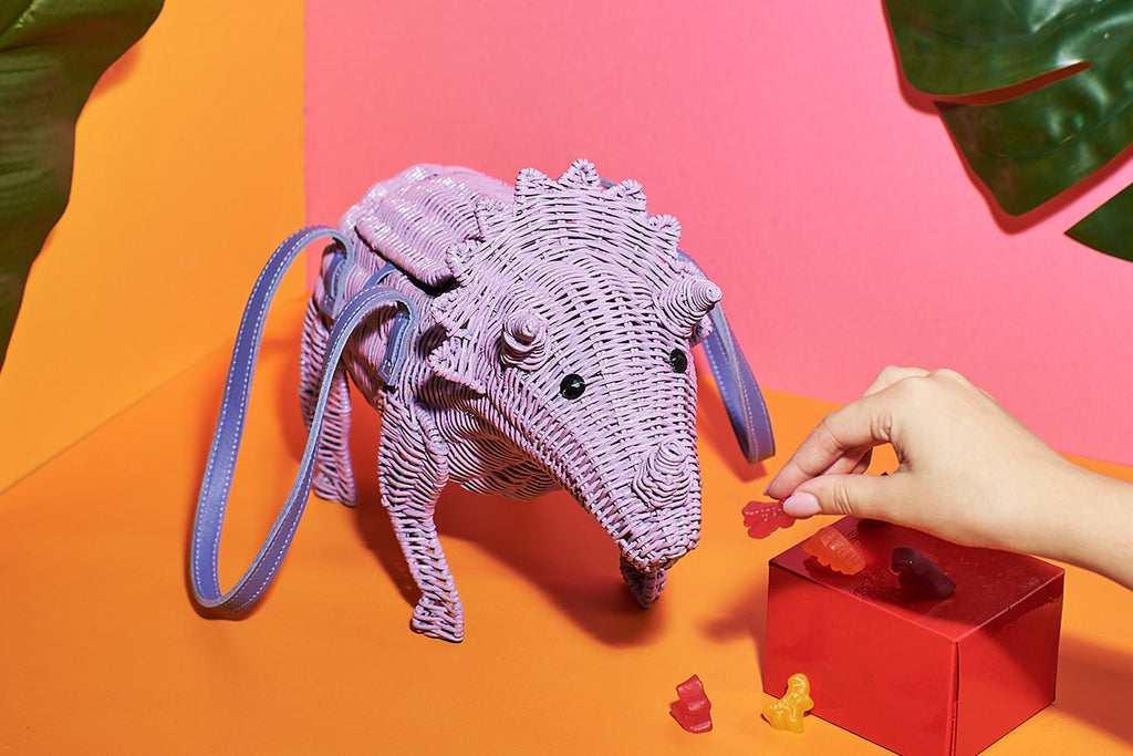Animal shaped Purse The purple coloured Patricia the Triceratops handbag by Wicker Darling sits in front of a pink and orange wall. The large wicker handbag is being fed dinosaur shaped gummies for a playful aesthetic.