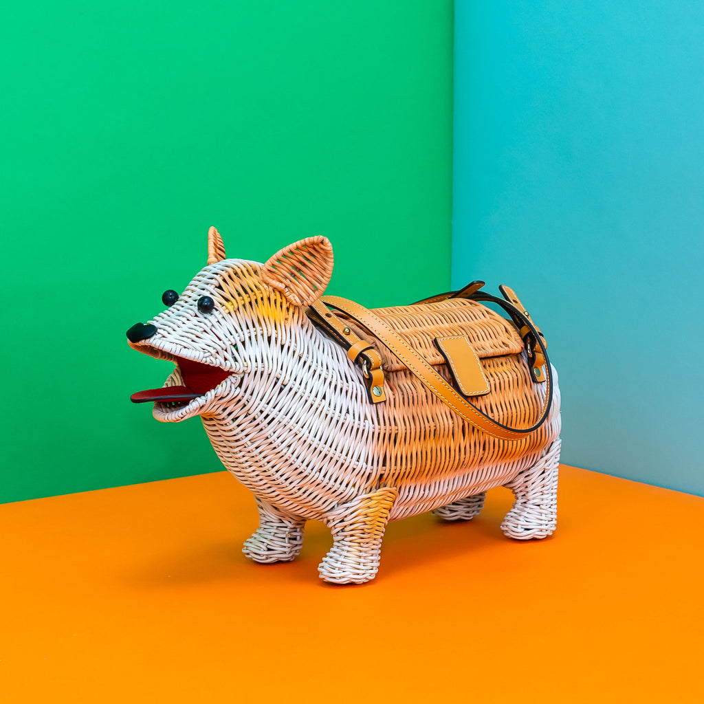 A rattan woven corgi-shaped bag. It's white and tan, with leather straps, big black bead eyes, a heart shaped nose, and the mouth is open as if it's smiling.