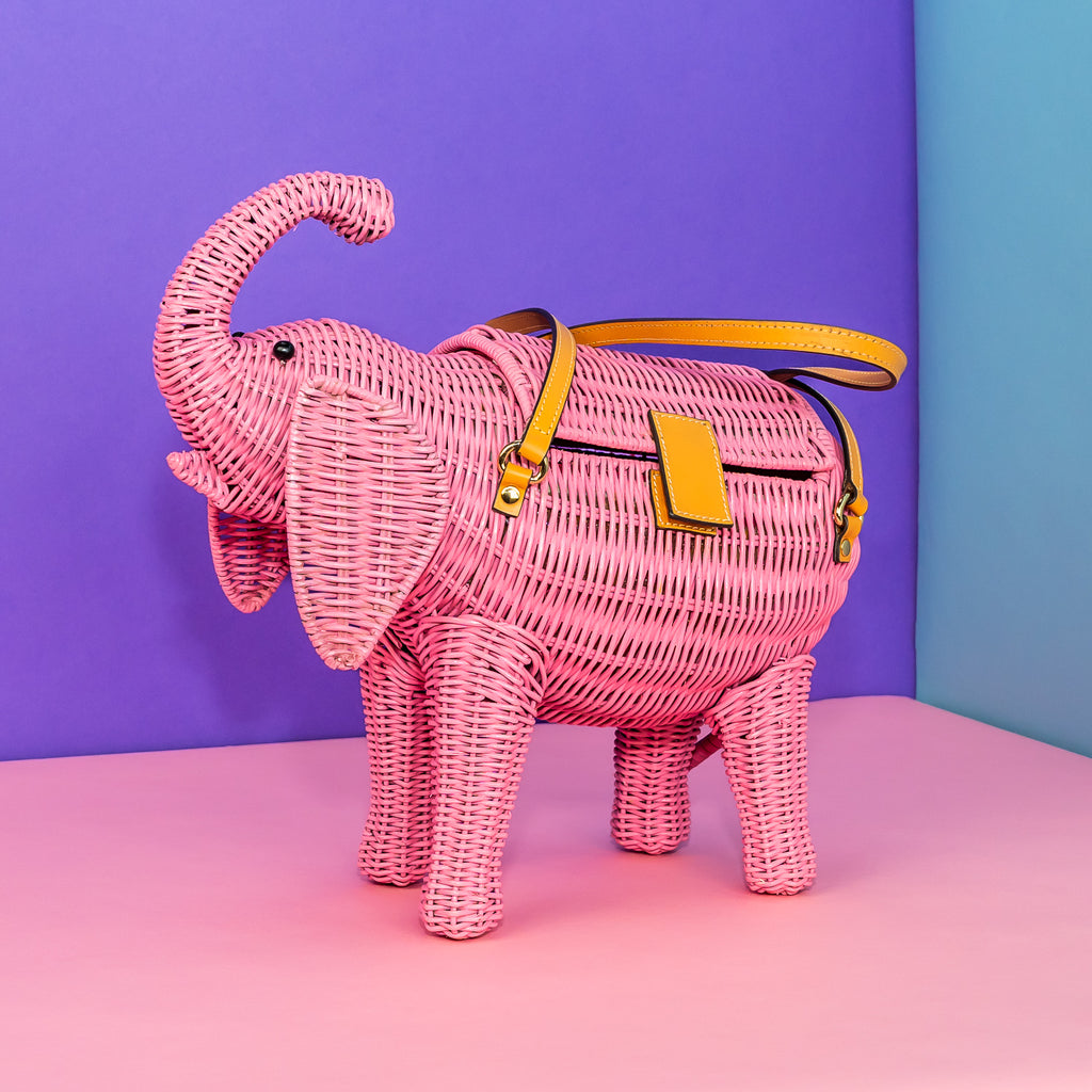 Wicker darling pink pachyderm elephant shaped handbag sits in a colourful background.
