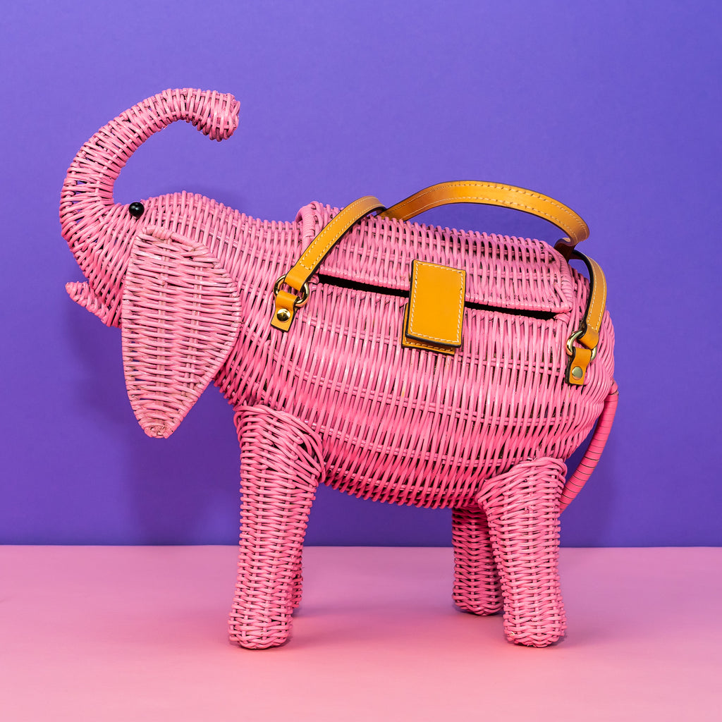 Wicker darling pink pachyderm elephant shaped handbag sits in a colourful background.