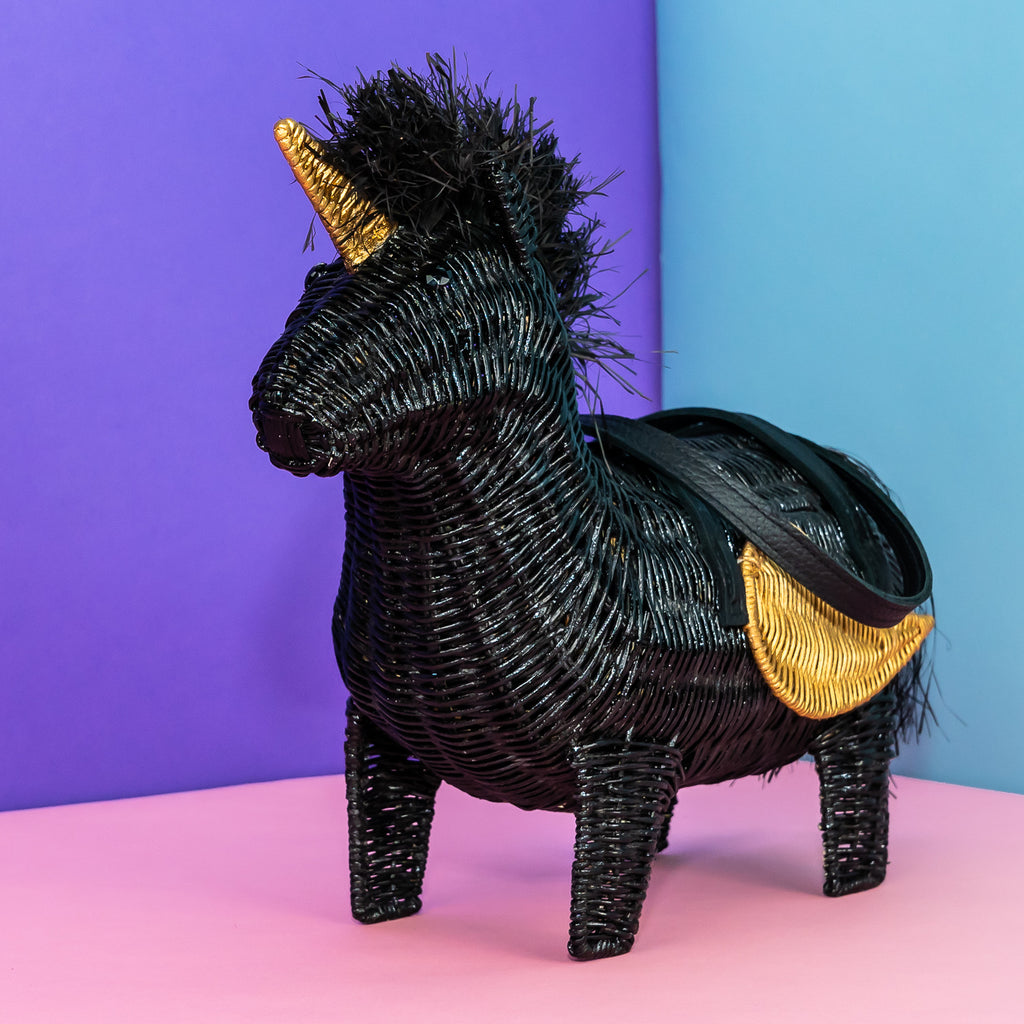 Wicker Darling vinny black unicorn bag with gold detailing sits in a colourful room.