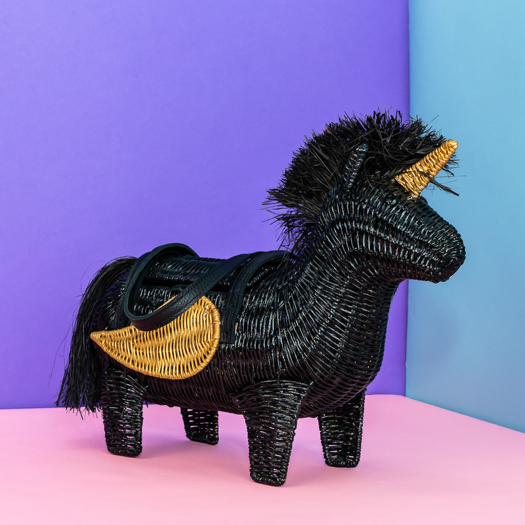 Wicker Darling vinny black unicorn bag with gold detailing sits in a colourful room.