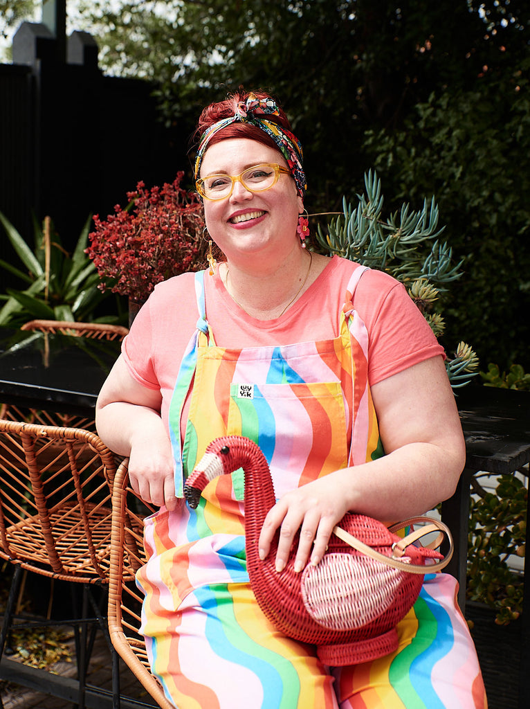Australian Designers Jasmine Norrie Wicker Darling founder sits with wicker handbag in her garden. she's wearing rainbow overalls with her handmade bags online Australia.