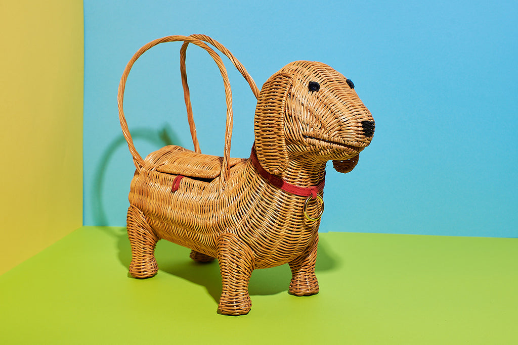 Animal Handbags A natural wicker coloured sausage dog handbag sits in a bright yellow, green and blue space. The bag features wicker handles and a red collar.