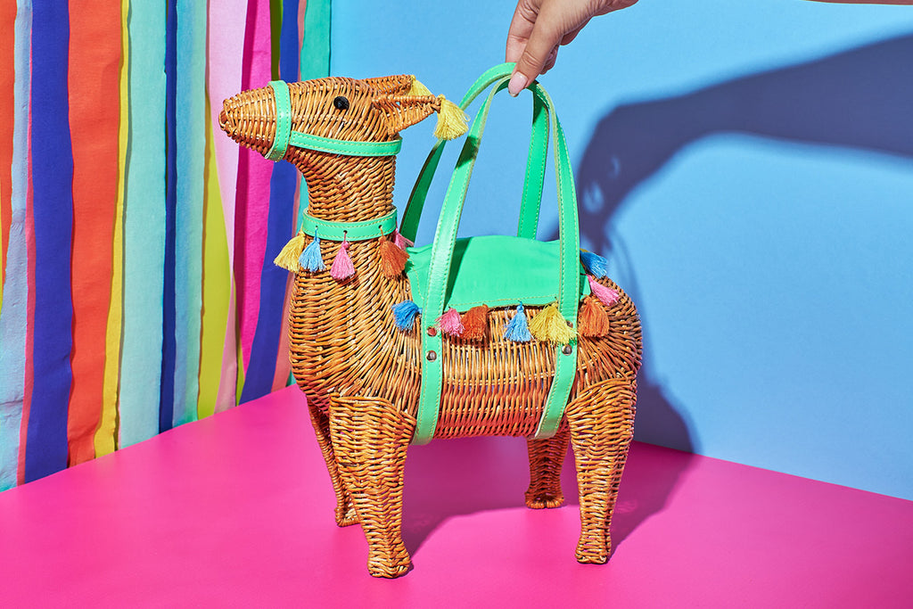 Animal Handbags A natural wicker coloured llama shaped handbag sits in front of a rainbow wall. The Wicker darling purse features green leather saddle and harness with rainbow pompoms around the edges. 