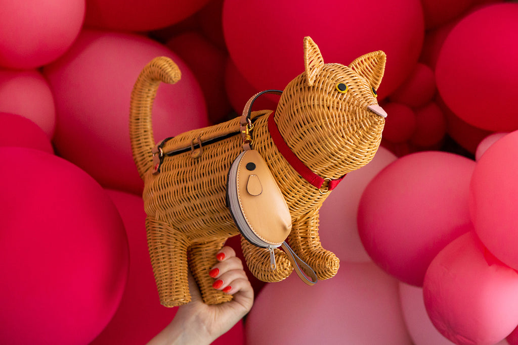 Orangey the orange tabby cat bag by Australian label Wicker Darling features a red bow with a tan coloured mouse-shaped purse hanging off of the colour. This image shows a hand with red nails holding Orangey in front of a red and pink balloon wall.