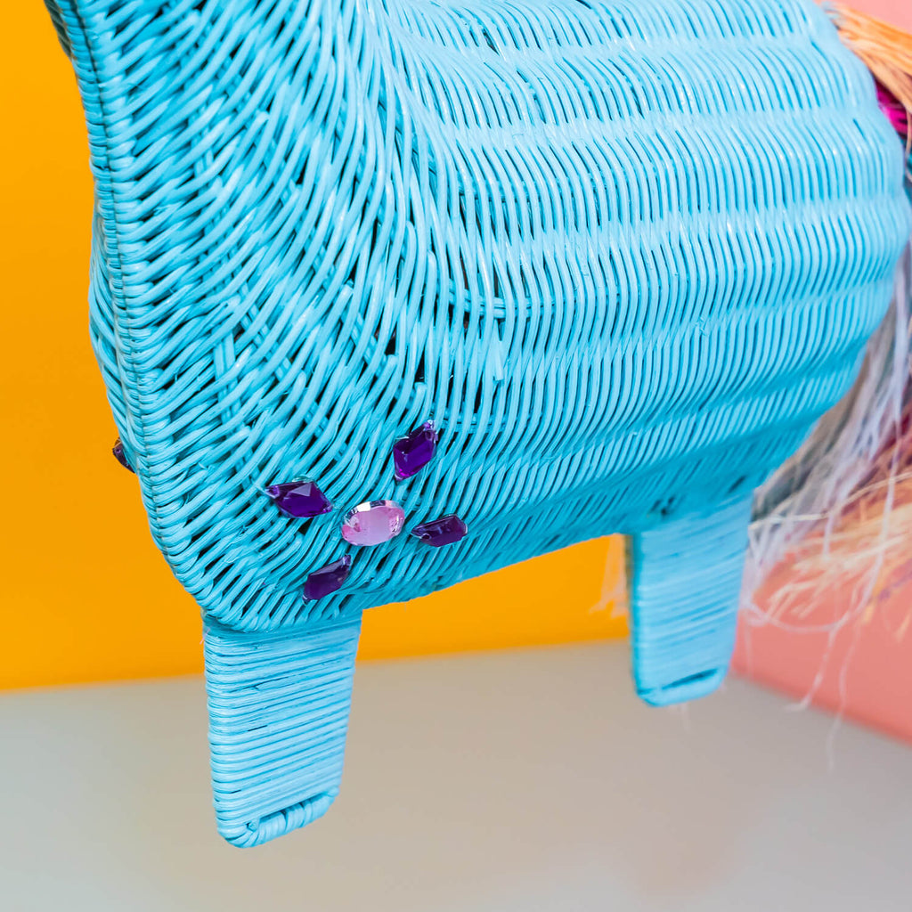 Wicker Darling aqua unicorn with multi-coloured pink rainbow main unicorn clutch bag is held in a colourful room. 