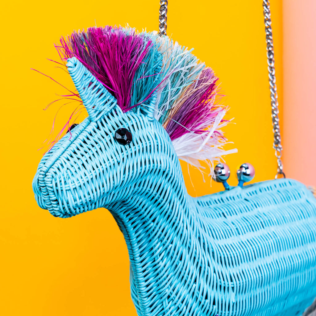 Wicker Darling aqua unicorn with multi-coloured pink rainbow main unicorn clutch bag is held in a colourful room. 