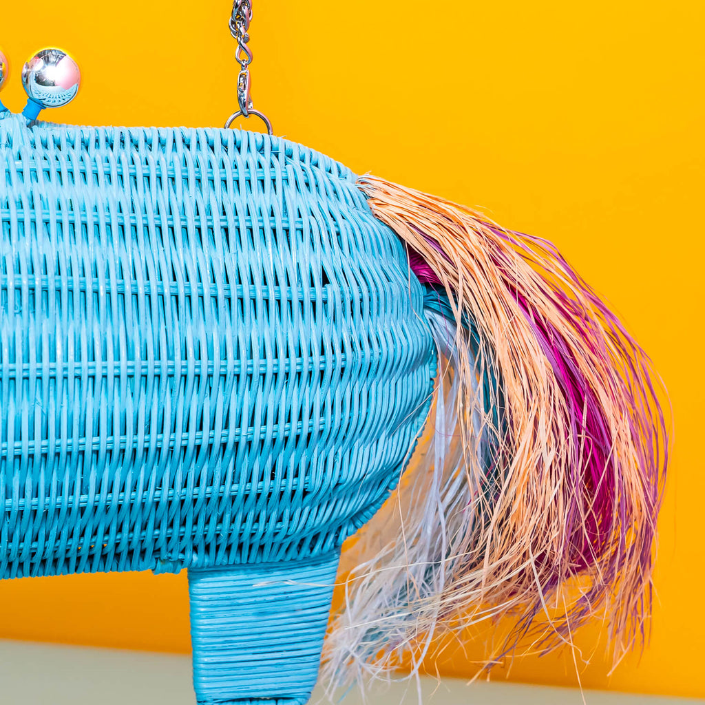 Wicker Darling aqua unicorn with multi-coloured pink rainbow main unicorn clutch bag is held in a colourful room. 