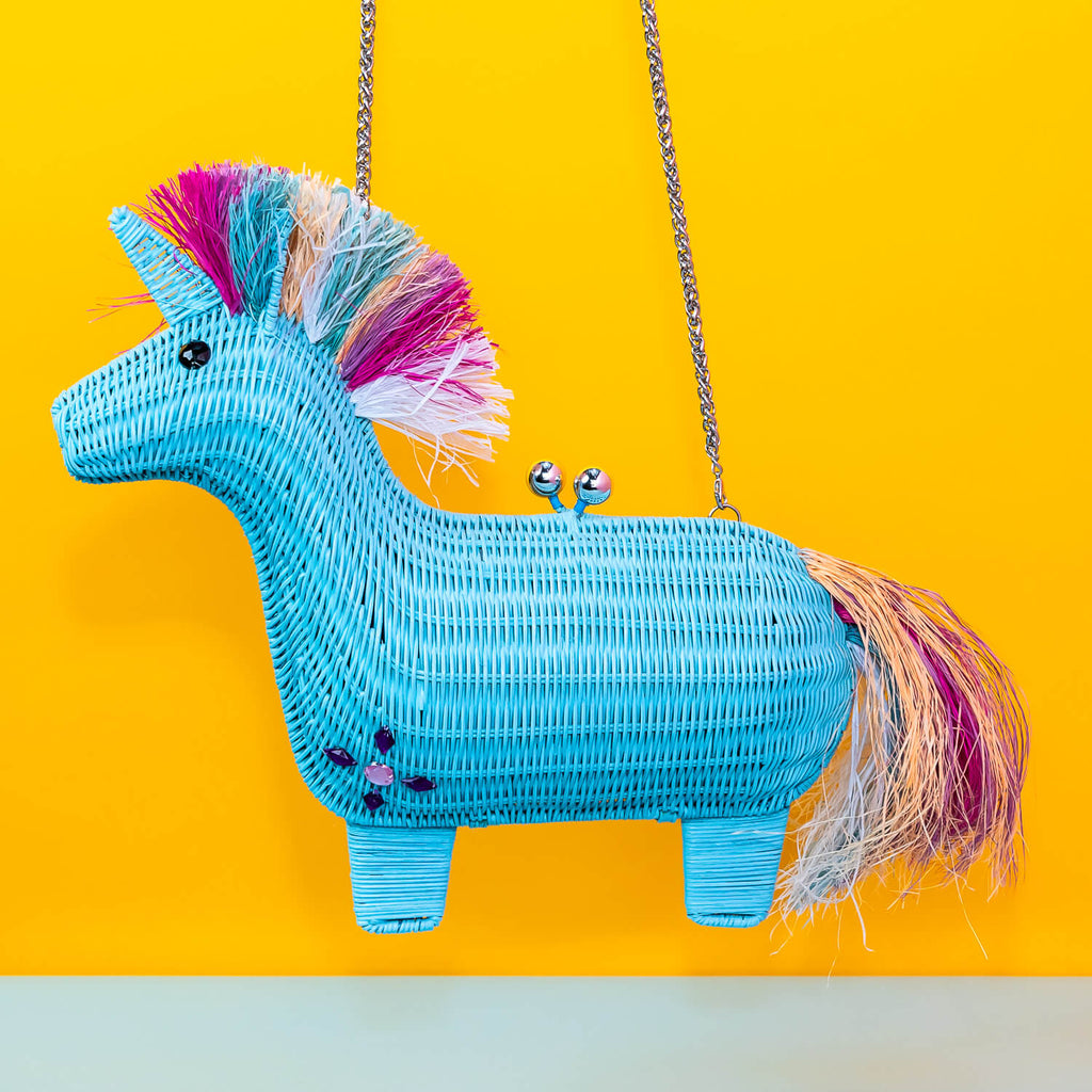 Wicker Darling aqua unicorn with multi-coloured pink rainbow main unicorn clutch bag is held in a colourful room. 