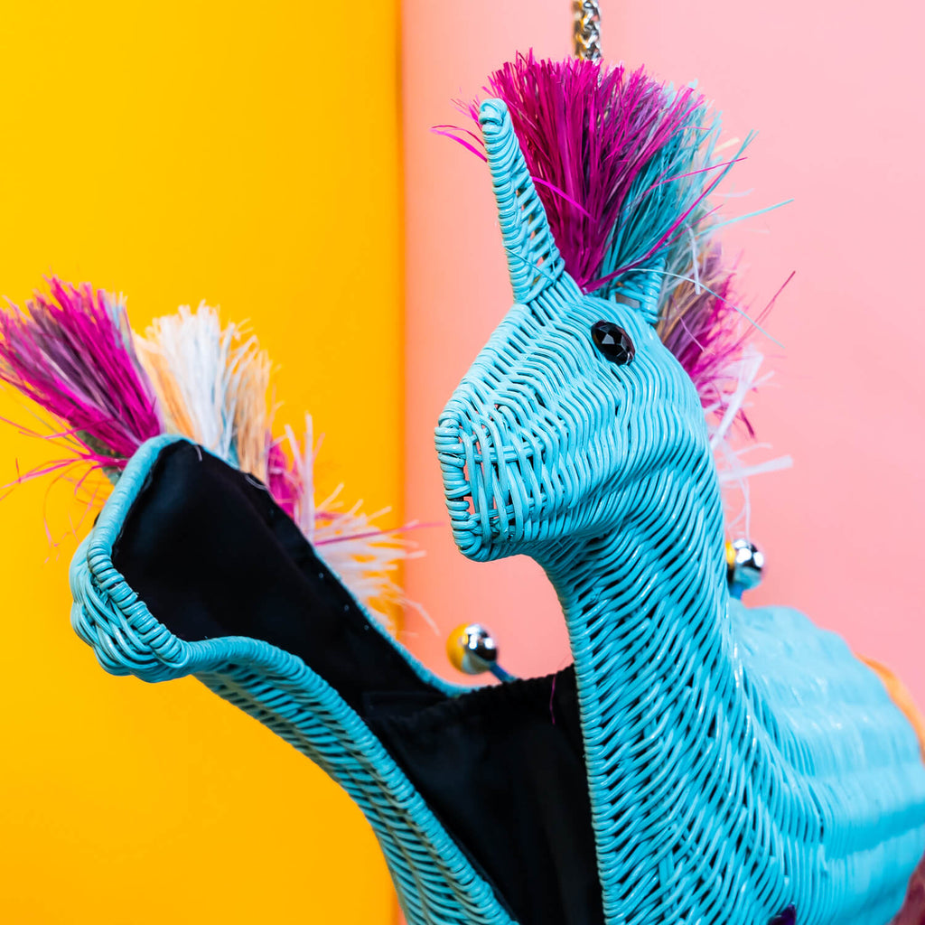 Wicker Darling aqua unicorn with multi-coloured pink rainbow main unicorn clutch bag is held in a colourful room. 