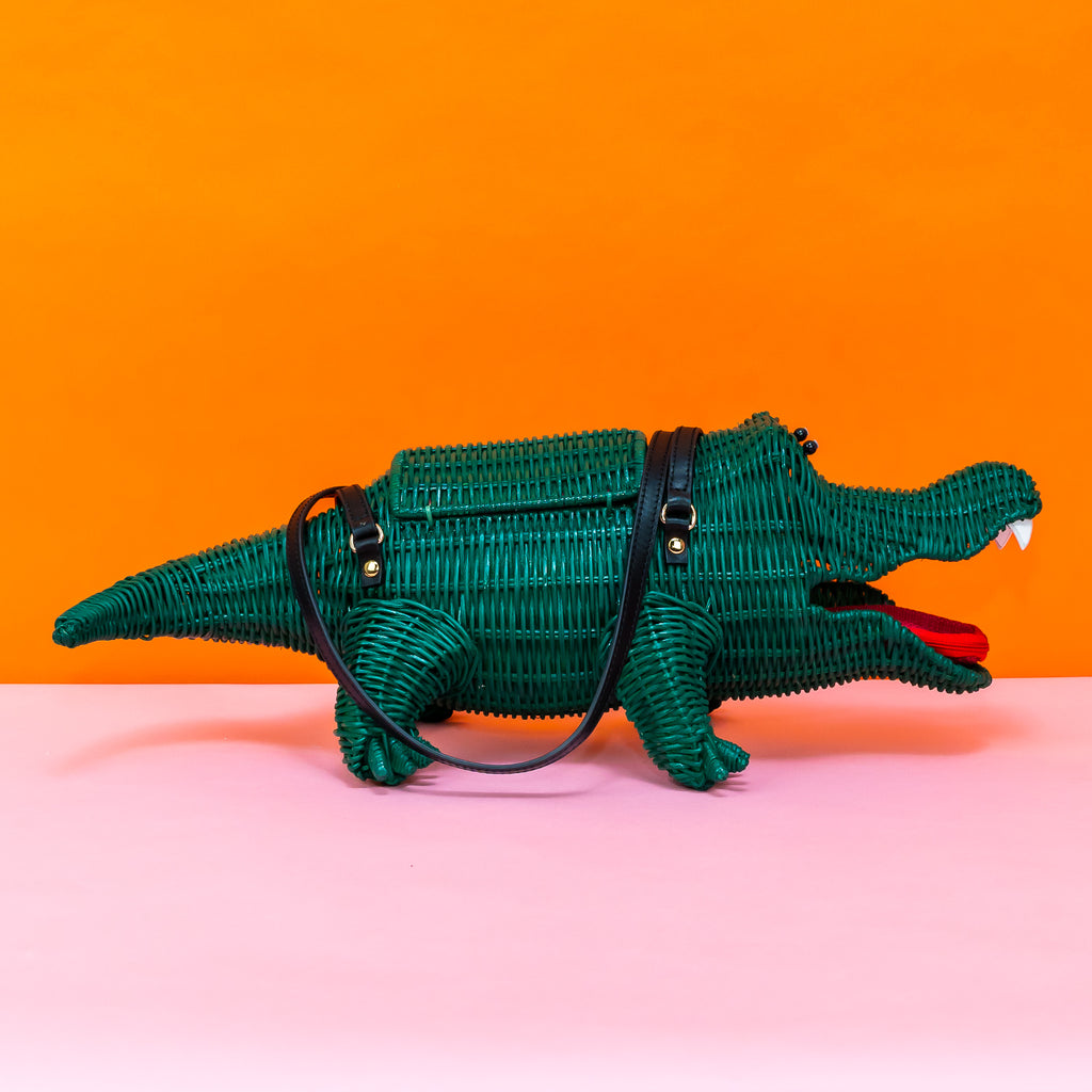 A profile/side view of Wicker Darling's green wicker crocodile handbag. It has black leather handles, and black bead eyes, as well as white leather teeth and a reddish pink coin purse tongue in the mouth of the bag.
