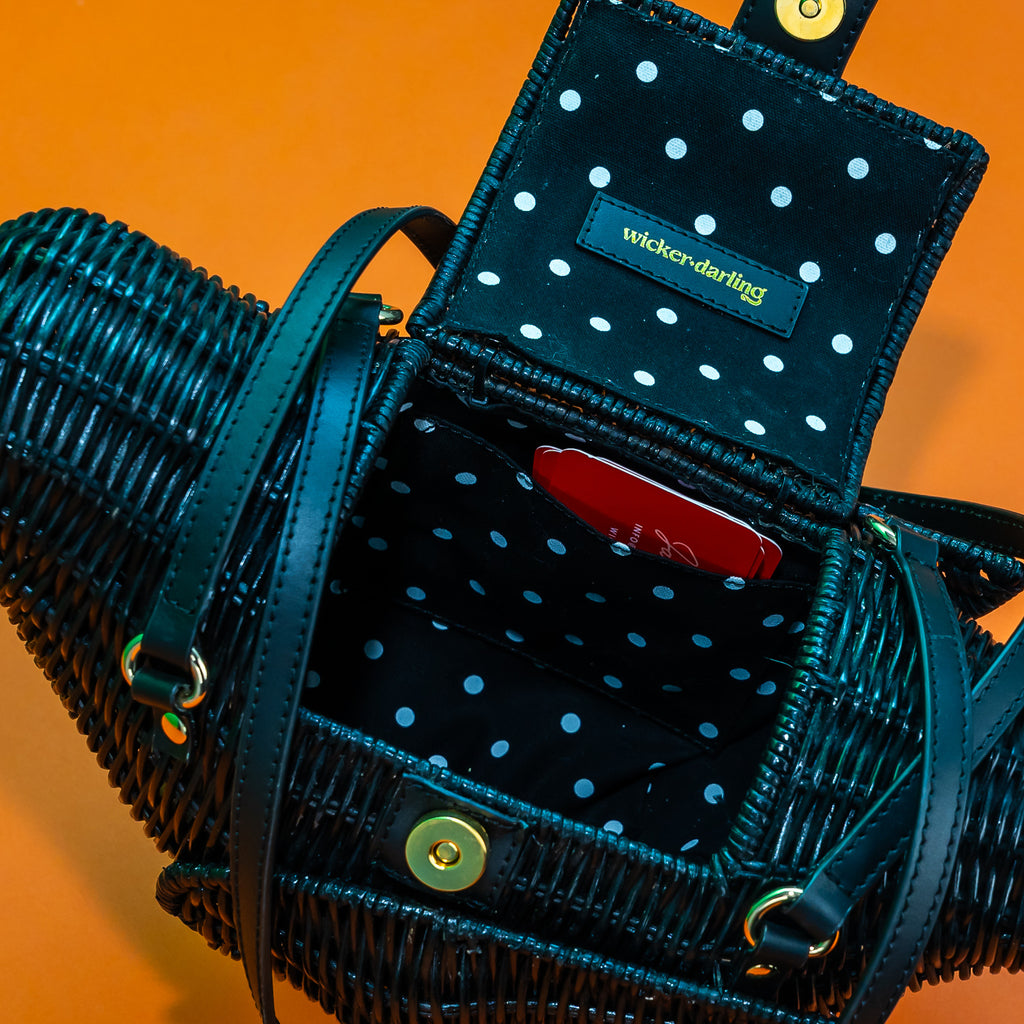 A close up view of the inside of a black rattan bird bag. The lining is a black and white pindot cotton. There's a pocket inside the lining, and a leather tag with 'Wicker Darling' stamped in gold.