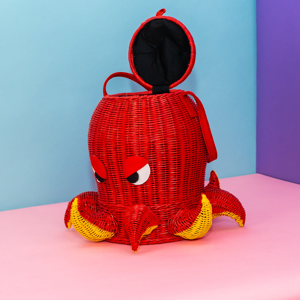 A woven rattan bag in the shape of an octopus. He is painted red, with yellow paint on the underside of his short tentacles. The tentacles alternate between up and down. He has leather eyes shaped to resemble a deep frown, and a single red strap to carry him by. His lid is open and shows a black lining.
