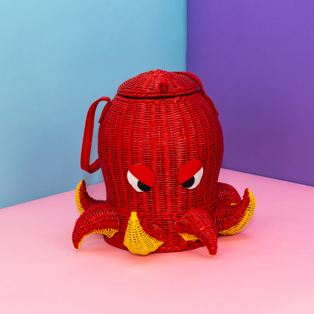 A woven rattan bag in the shape of an octopus. He is painted red, with yellow paint on the underside of his short tentacles. The tentacles alternate between up and down. He has leather eyes shaped to resemble a deep frown, and a single red strap to carry him by.