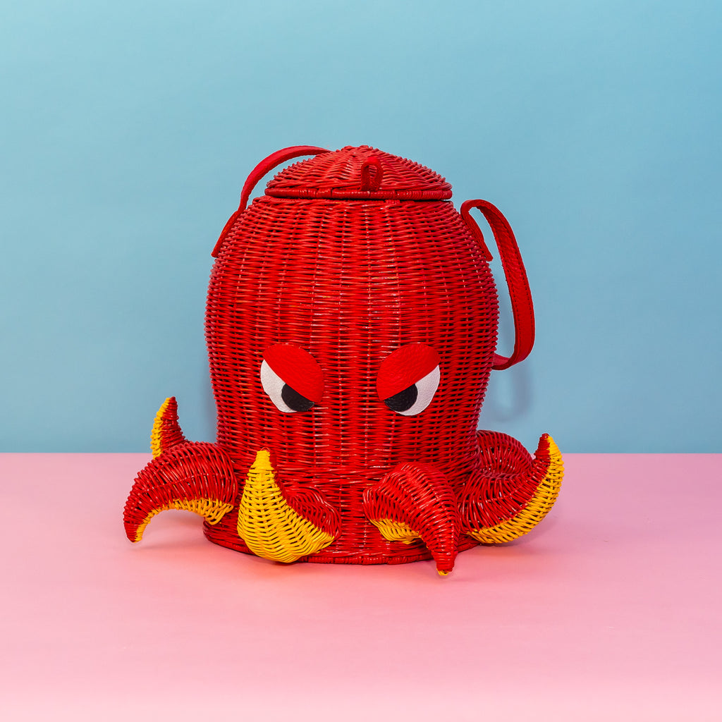 A woven rattan bag in the shape of an octopus. He is painted red, with yellow paint on the underside of his short tentacles. The tentacles alternate between up and down. He has leather eyes shaped to resemble a deep frown, and a single red strap to carry him by.