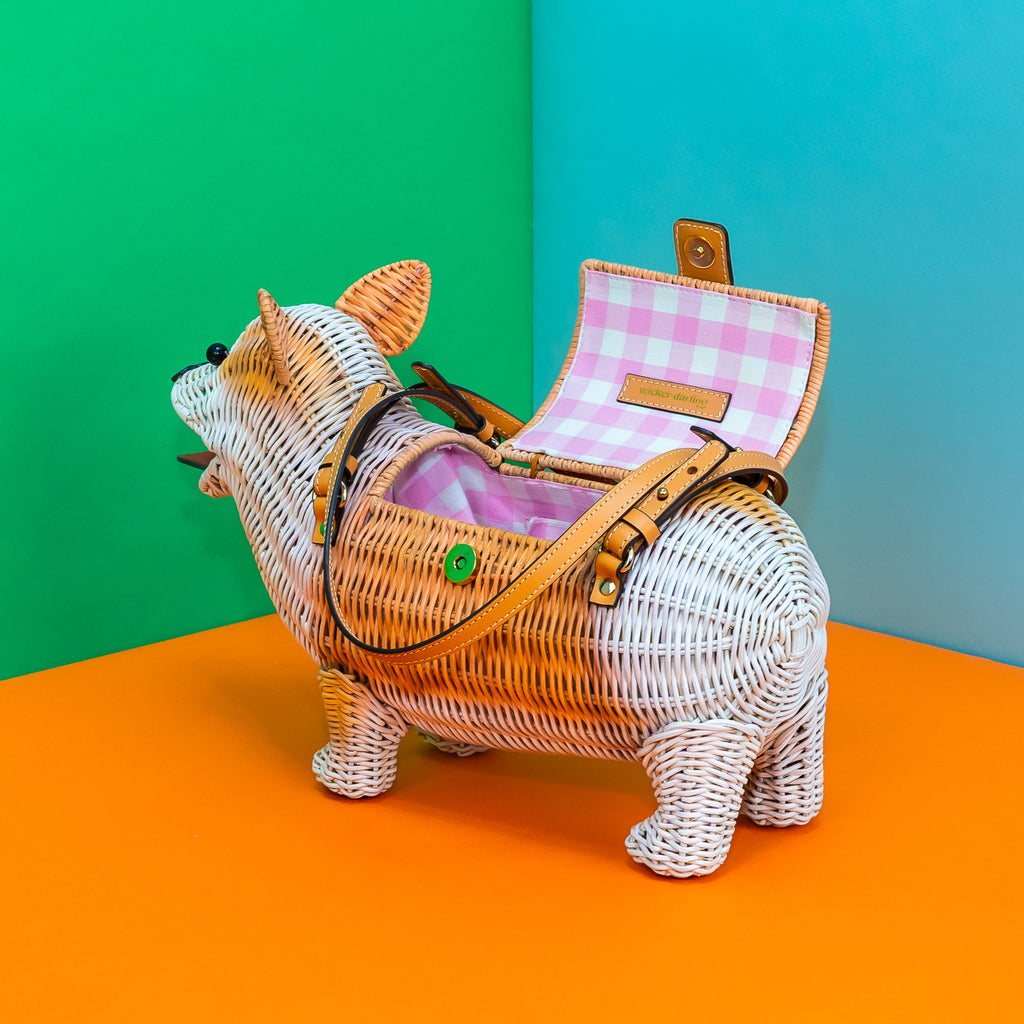 A side-to-back view of Wicker Darling's rattan corgi bag. It has a white bottom and white legs; the lid is open revealing a pink gingham lining.