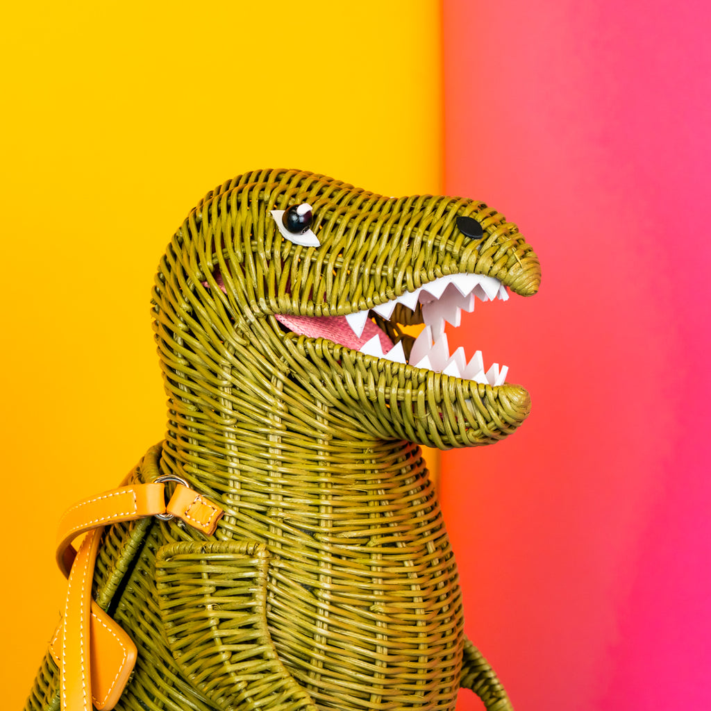 A green t-rex wicker bag with open mouth full of white leather teeth and pink tongue.