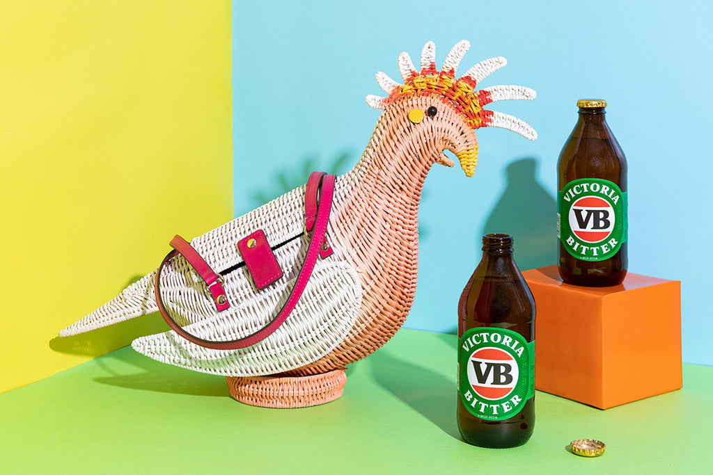 A major mitchell shaped wicker handbag by Wicker Darling sits in front of a yellow, green and blue background. The pink and yellow parrot bag features pink handles and is sitting in front of two VB beers for a true Australiana aesthetic.