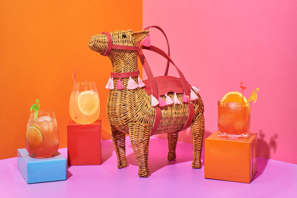 Animal Handbags A natural wicker coloured llama shaped handbag sits in front of an orange, pink and purple wall. The Wicker darling purse features pink leather saddle and harness with various pink pompoms around the edges.