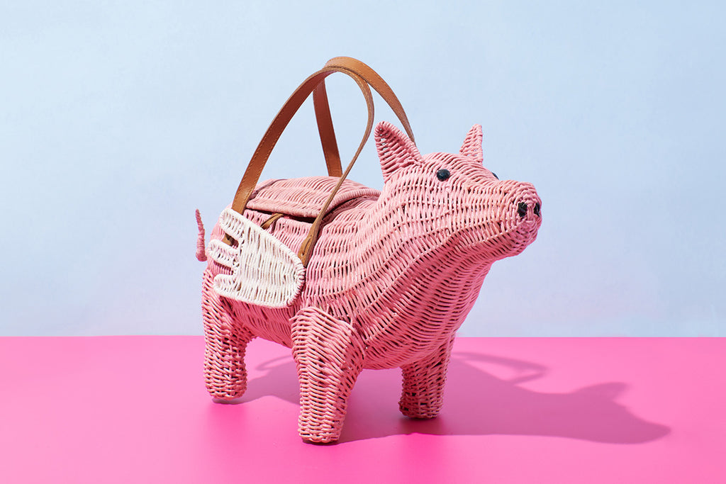 A pink pig shaped purse with white wings is fashioned into a wicker handbag by Australian fashion designer Wicker Darling. The bag has a tan leather handle and sits in front of a pink and blue background.