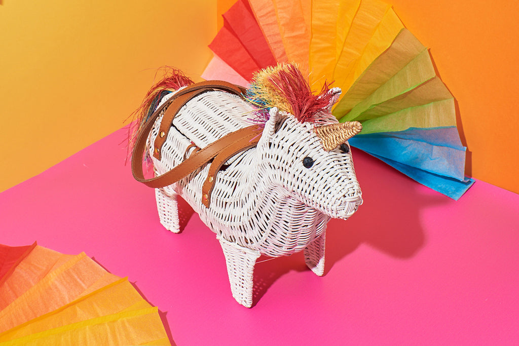 A white unicorn with a rainbow mane by Australian designer Wicker Darling sits in front of a pink and yellow wall. The creature has a yellow horn and the quirky purse features tan coloured leather handles.