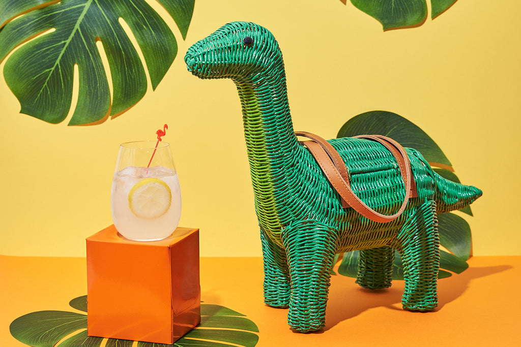 Animal Handbags The Charlotte Brontesaurus dinosaur shaped handbag by Wicker Darling sits in an orange and yellow wall with leaves. The green bag with brown handles has a cocktail in front of it to match the retro party scene.
