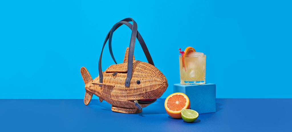 Unique Purses Wicker Darling Marcus the sharkus shark purse sits with a cocktail in front of a blue background