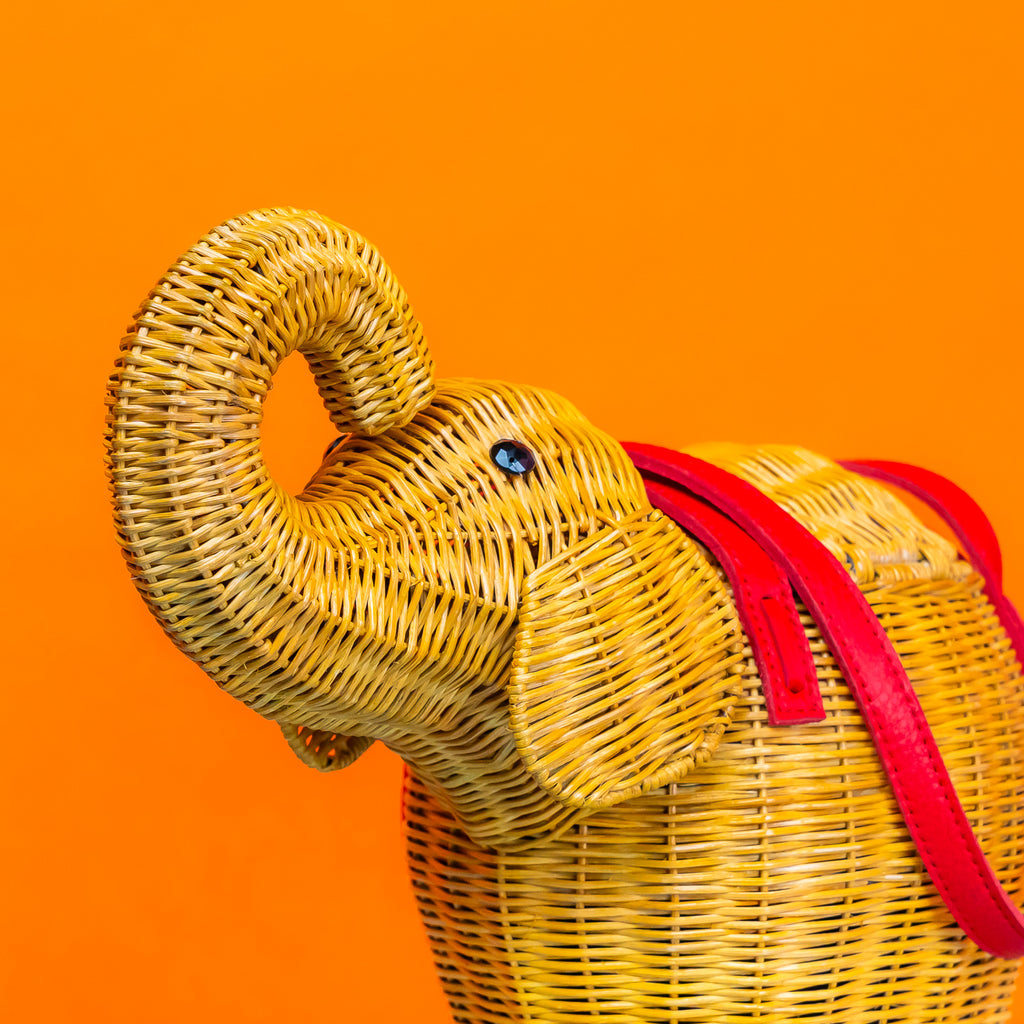 A close up of the trunk and head of a rattan wicker bag shaped like an elephant, with red leather handles.