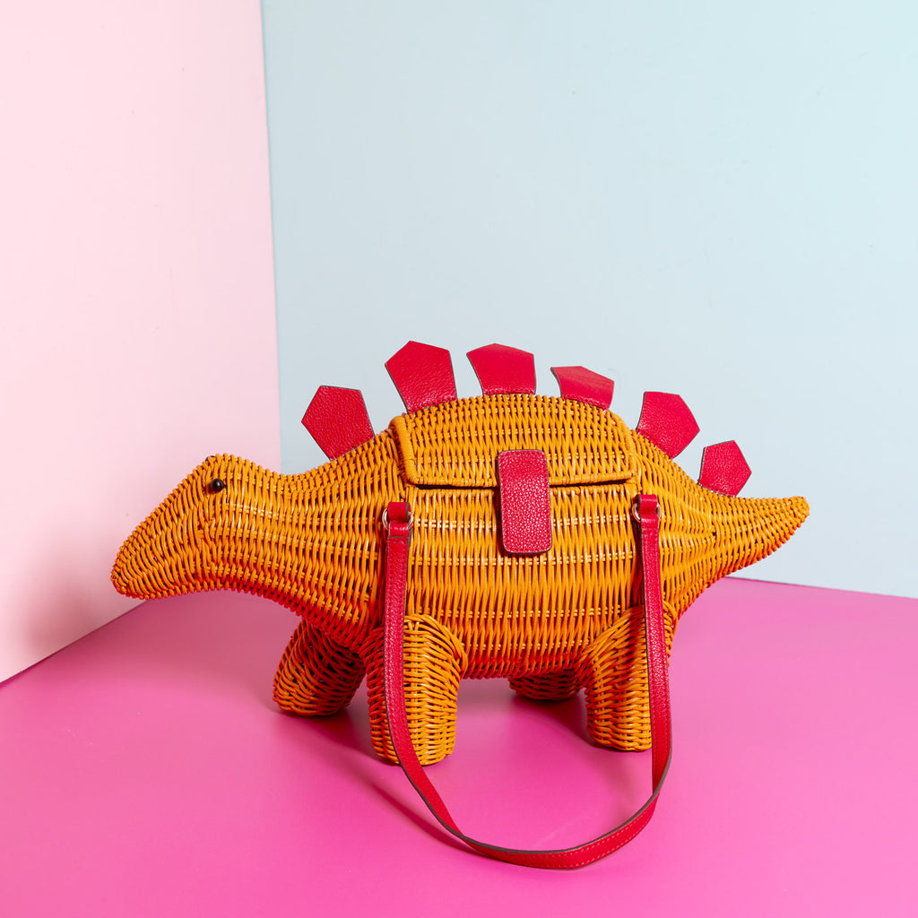 Wicker Darling stegosaurus bag is bright yellow with red leather detailing sits in a colourful background