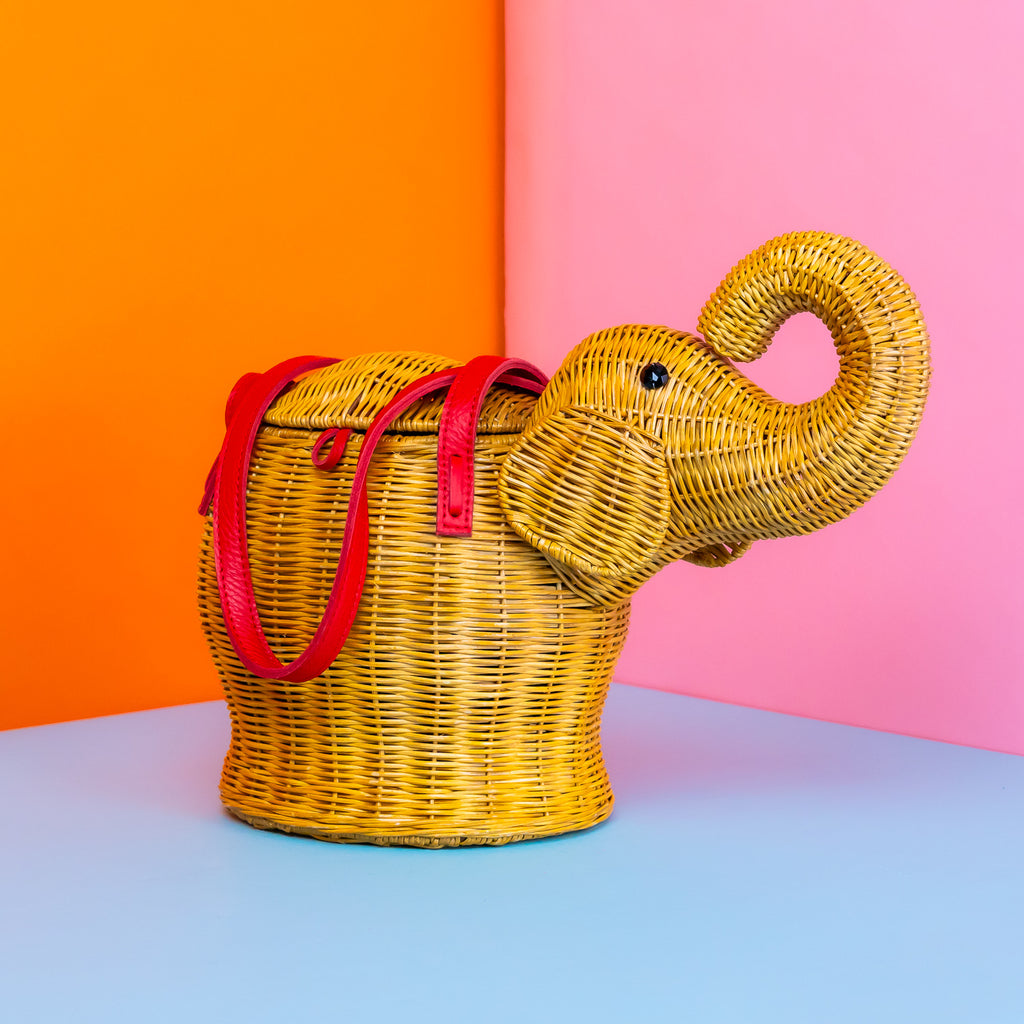 A photo of a rattan wicker bag in the shape of an elephant. it has its trunk curled up, and red leather handles.