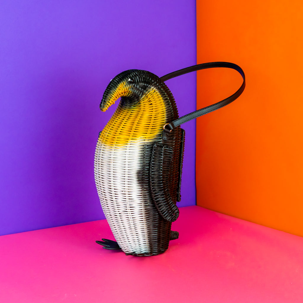 Penguin-shaped king penguin handbag on a colorful background. The penguin is standing upright and has two leather feet, a black leather elbow strap, and the body is rattan painted black, yellow and white with king penguin markings. 