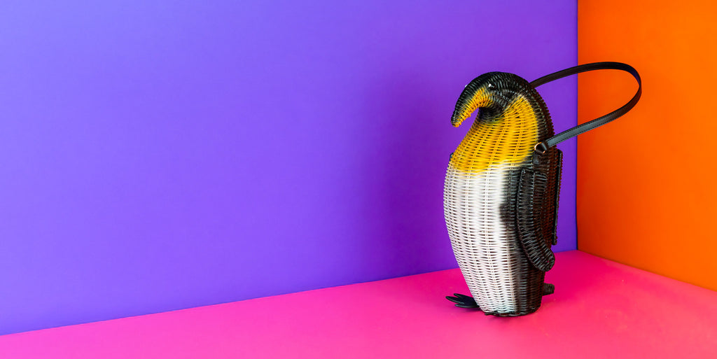 Penguin-shaped king penguin handbag on a colorful background. The penguin is standing upright and has two leather feet, a black leather elbow strap, and the body is rattan painted black, yellow and white with king penguin markings. 
