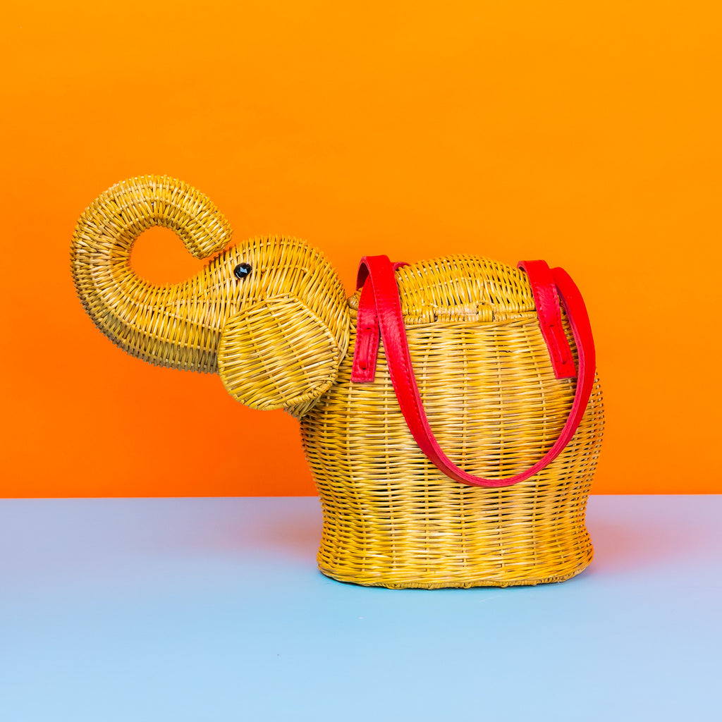 An elephant shaped bag with the trunk up; the elephant is in natural rattan with red leather handles.