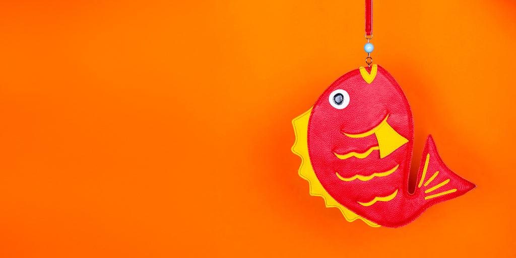 Red fish shaped leather wristlet fish shaped bag with yellow accents hangs in front of an orange background