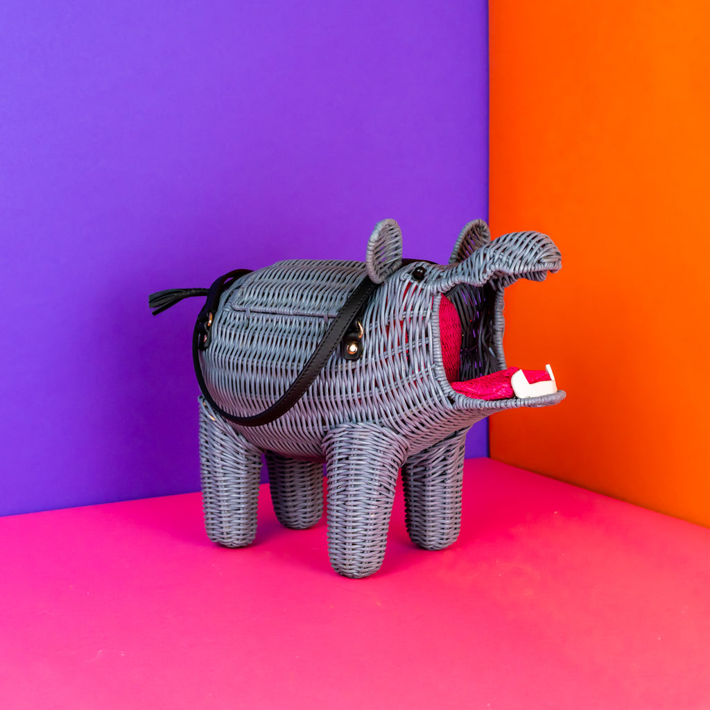 Wicker Darling chunky wicker hippo purse in medium grey with black leather straps. The mouth is wide open and reveals a bottom row of white leather teeth, and a pink fabric tongue with zipper down the middle, revealing a coin purse hidden in the tongue. 