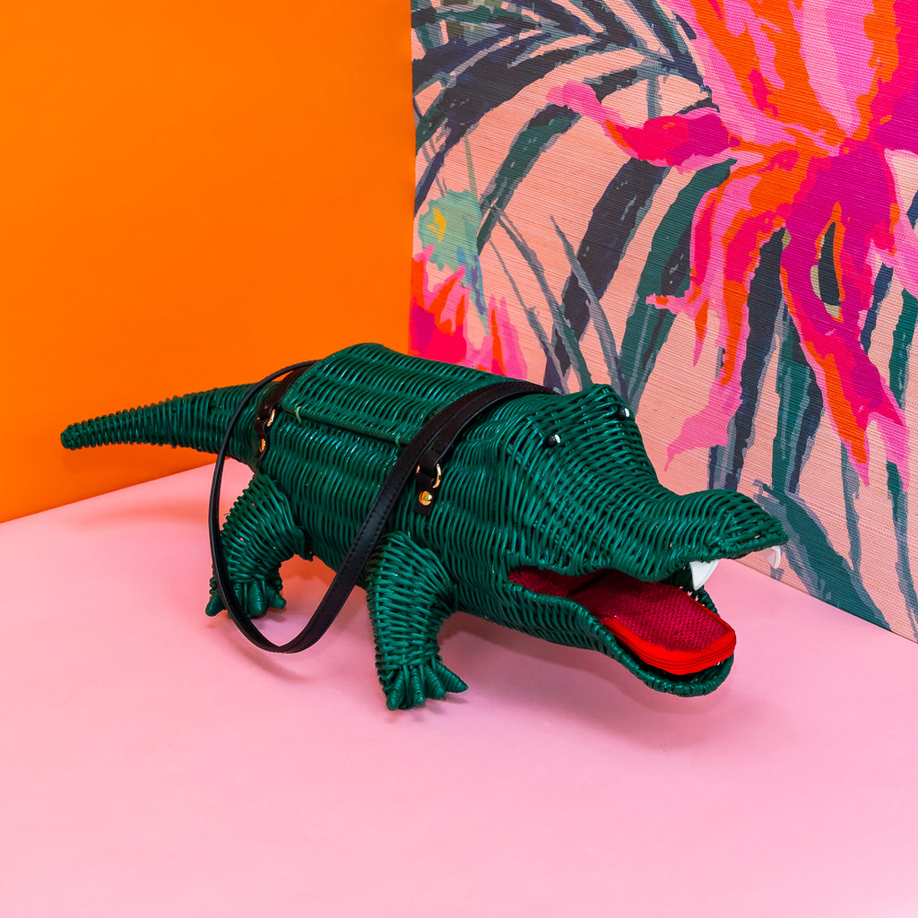 A rattan handbag shaped like a crocodile, painted green, with black leather handles. It has white leather teeth, and a dark pinky-red tongue that doubles as a zippered coin purse.