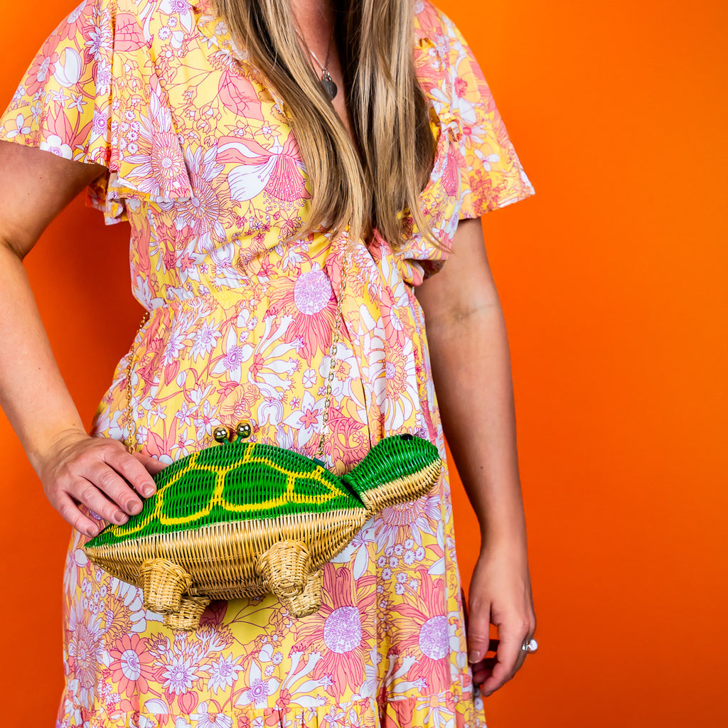 Shelby turtle shaped bag turtle handbag is worn by a woman in a floral dress in front of an orange background.