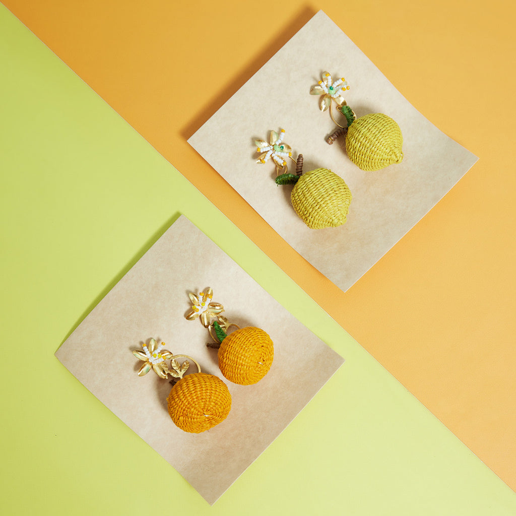 wicker darling hand woven citrus stud earrings in the shape of lemons and oranges sit in a colourful background