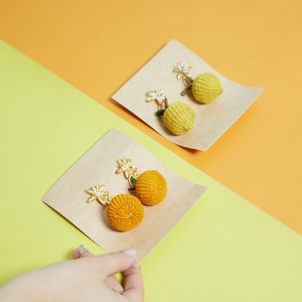 wicker darling hand woven citrus stud earrings in the shape of lemons and oranges sit in a colourful background